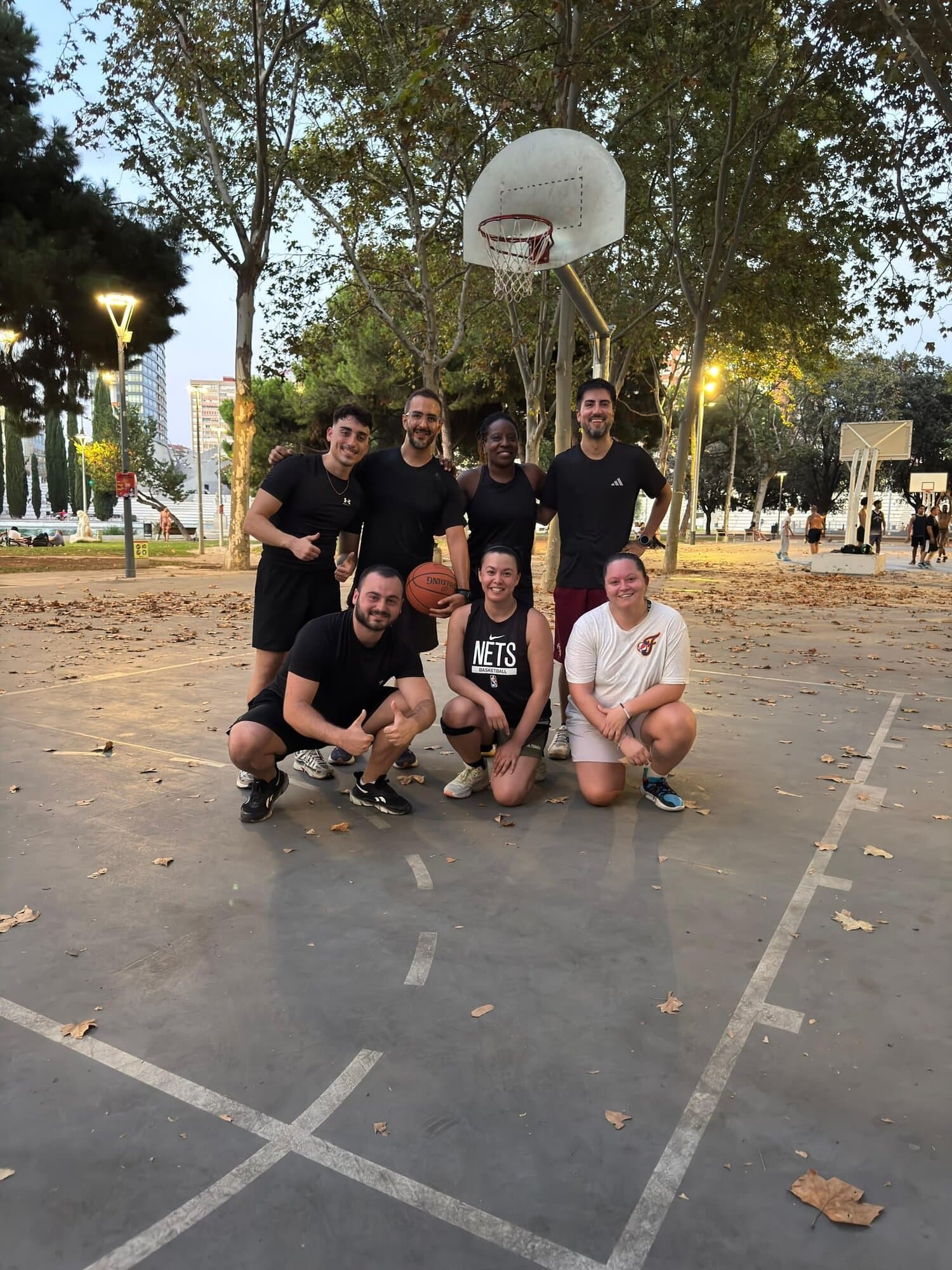 Mixed group of Keep Calm Community Barcelona members posing with a basketball on an outdoor public court after a pickup game