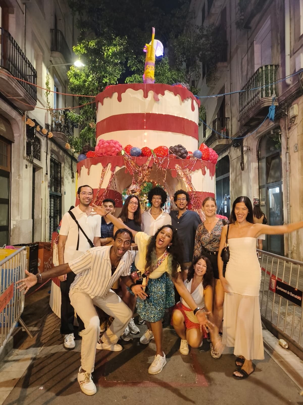 Keep Calm Community Barcelona members posing with arms wide in front of a giant cake decoration at a night-time street festival
