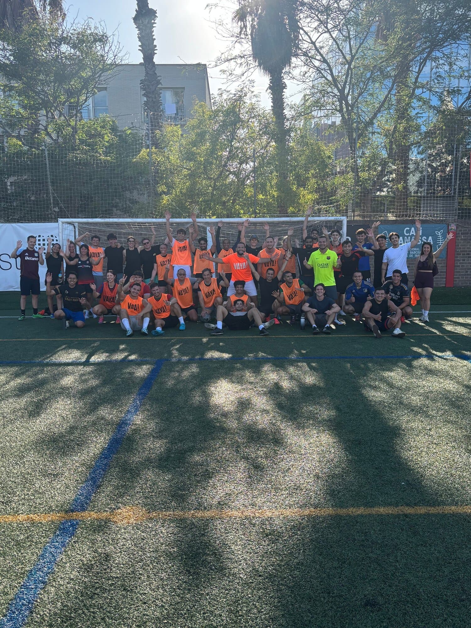 Large Keep Calm Community Barcelona football group photo, players in orange Vaya Sports bibs celebrating together after back-to-back Friday games on an outdoor pitch