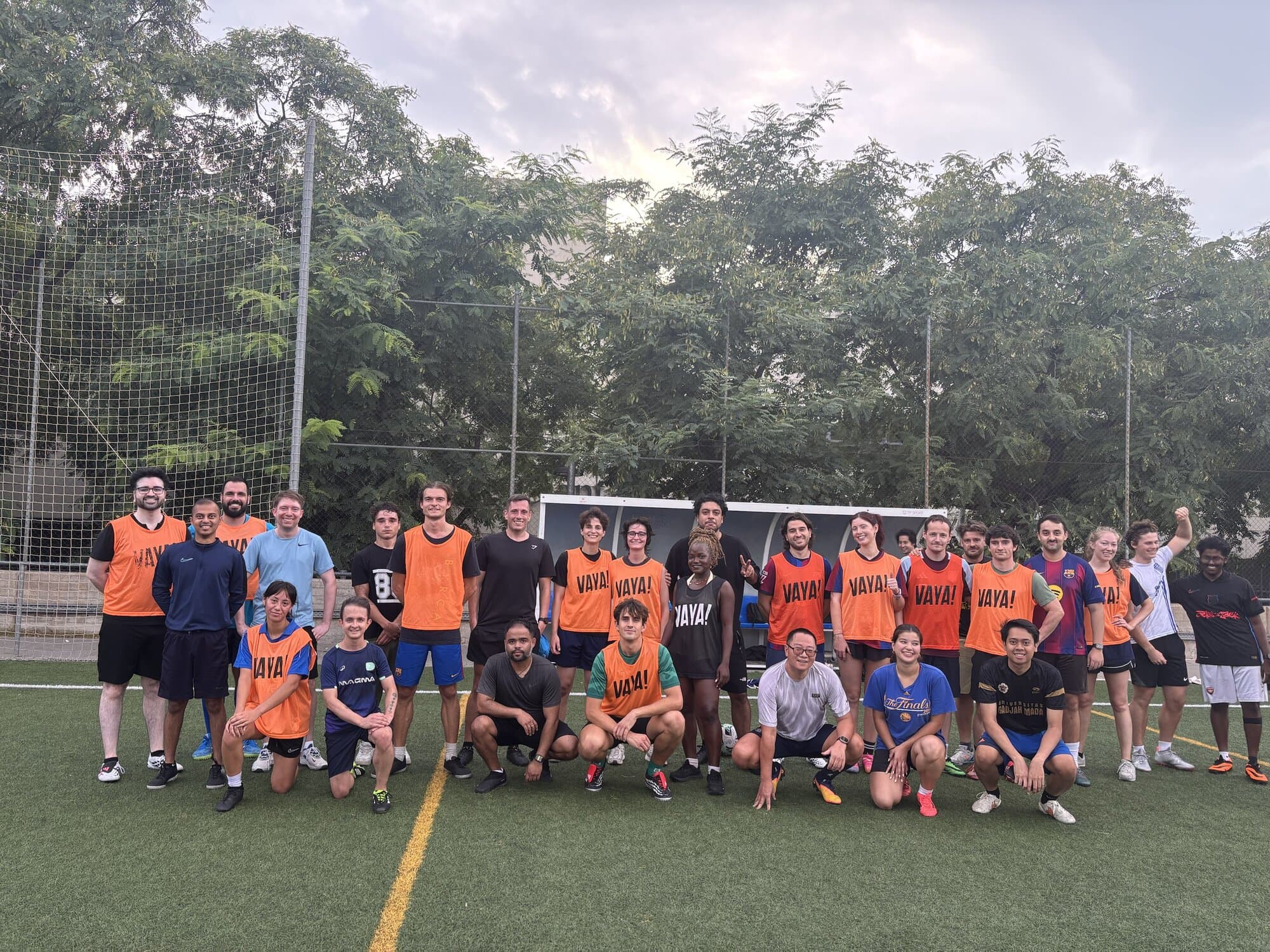 Keep Calm Community Barcelona football group photo, players in orange Vaya Sports bibs posing together on an outdoor turf pitch at dusk after a Thursday evening game