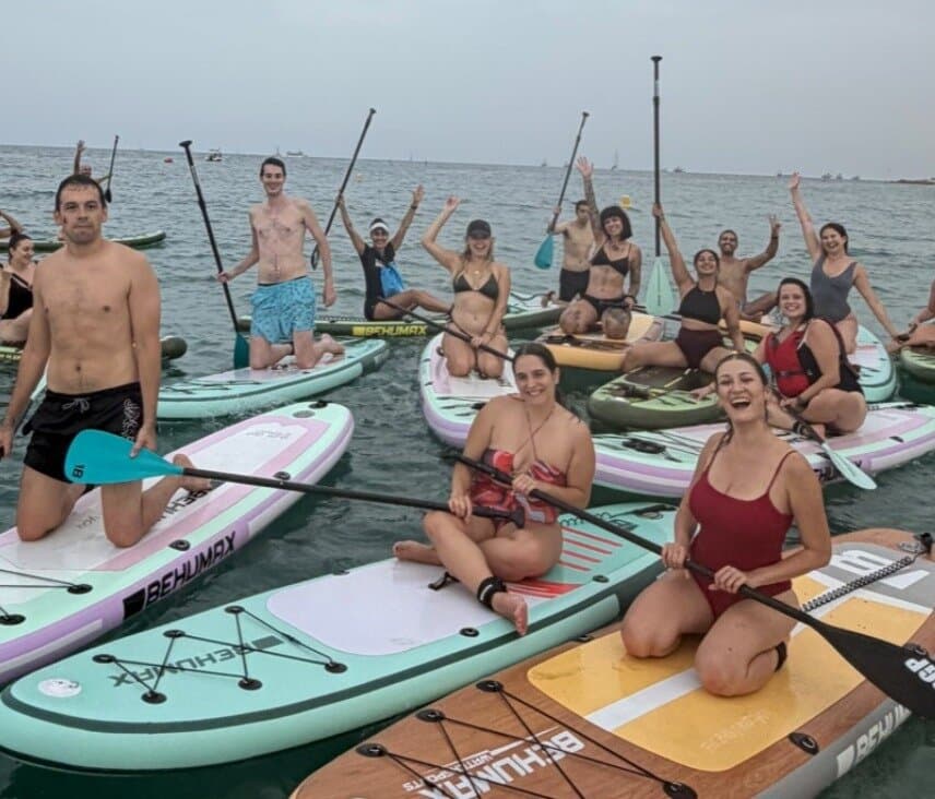 A large group of Keep Calm Community Barcelona members kneeling on paddleboards in the sea off the Barcelona coast, smiling and waving with paddles raised